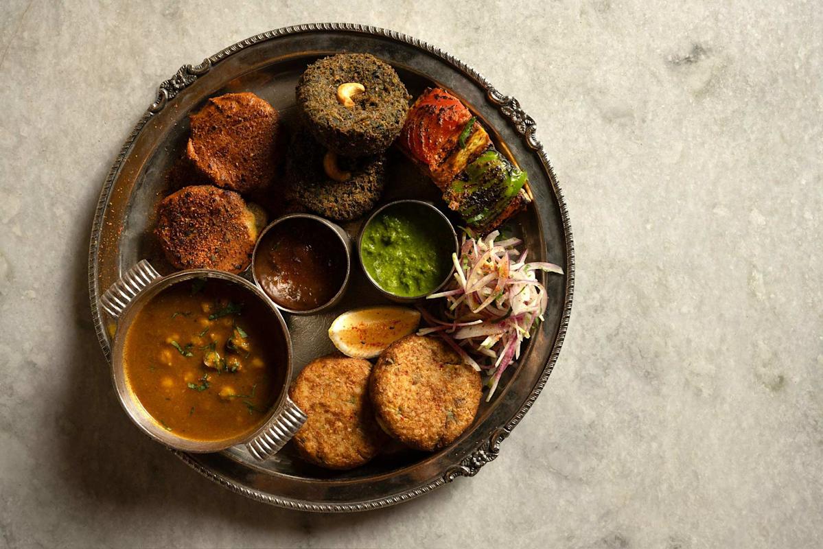 A traditional Indian thali on a metal plate featuring cutlets, kebabs, green chutney, dal in a bowl, grilled vegetables, sliced onions, a lemon wedge, and two small dipping sauces on a marble surface.