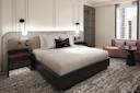 A modern hotel room with a large bed, white bedding, black and gold accent pillow, two bedside tables with lamps, a textured carpet, a mauve sofa, a round table, and large windows with city views.