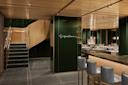 Upscale restaurant interior with green walls, a wooden ceiling, grey tiled floors, blue cushioned bar stools, neatly set tables, and a staircase with wooden handrails. Elegant lighting creates a warm, inviting atmosphere.