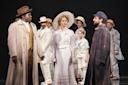 A group of actors in period costumes stand on stage; a woman in a white dress and hat faces two men, one in a beige coat and hat, the other in a dark coat and cap, with others in white attire watching behind them.