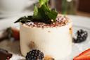 A creamy dessert topped with cocoa powder and fresh mint leaves is served on a plate. It is surrounded by blackberries and a sliced strawberry, with a dusting of powdered sugar.