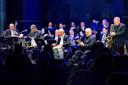 A large jazz band performs on stage, with musicians playing various instruments like saxophones, trumpets, and percussion. They are dressed in formal attire, creating a lively atmosphere under blue and purple stage lighting.