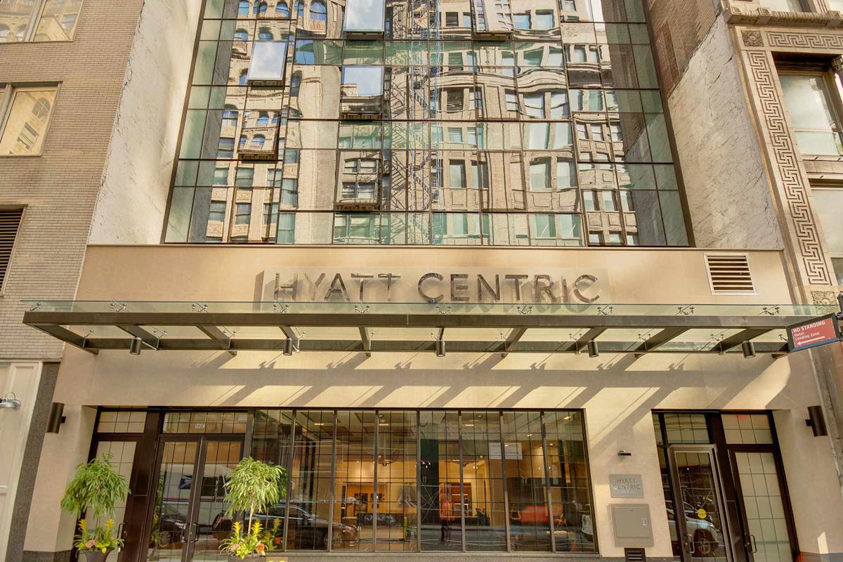 The entrance of a Hyatt Centric hotel with large glass windows reflecting nearby buildings; glass canopy and potted plants are visible at the entrance.
