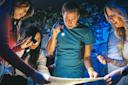 Four people are gathered around a table, looking intently at a map or clue, using small flashlights in a dimly lit room, suggesting they are participating in an escape room or puzzle-solving activity.