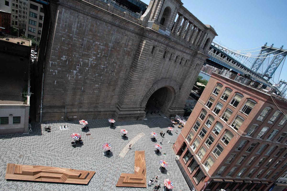 Aerial view of a plaza with people sitting at tables with red-and-white umbrellas, surrounded by tall buildings. A large stone archway and part of a suspension bridge are visible in the background.