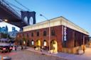 A brick building with a glowing rooftop, neon "THEATER" sign, and arched doorways sits beneath the Brooklyn Bridge at dusk, with people walking along the cobblestone street. Manhattan skyline is visible in the background.