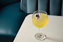 A yellow cocktail in a stemmed glass, garnished with a purple and white edible flower, sits on a white marble table beside a teal upholstered seat.