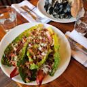 A plate of fresh romaine lettuce salad topped with bacon, onions, and crumbled cheese. In the background, a bowl of mussels with bread, glasses of water, and napkins with utensils on a wooden table.