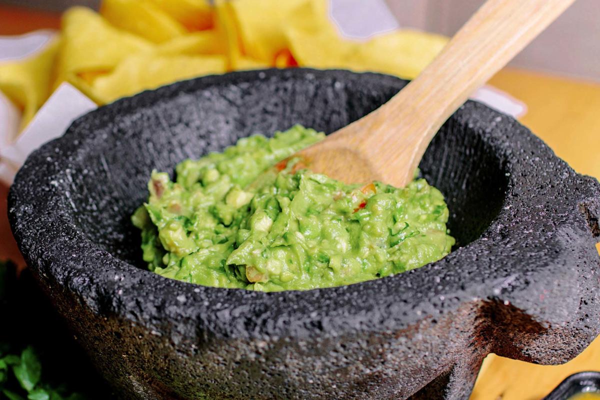A wooden spoon stirs fresh guacamole in a traditional black stone mortar, with a yellow cloth in the background.