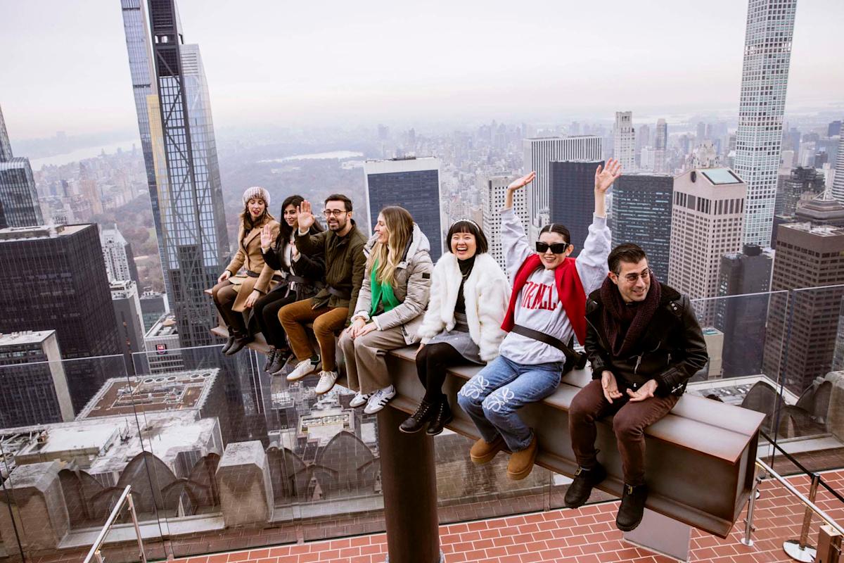 A group of people sitting on the edge of a high rooftop, with a sprawling cityscape in the background. They are smiling and waving, enjoying the scenic view of skyscrapers on a cloudy day.