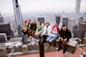 A group of people sitting on the edge of a high rooftop, with a sprawling cityscape in the background. They are smiling and waving, enjoying the scenic view of skyscrapers on a cloudy day.