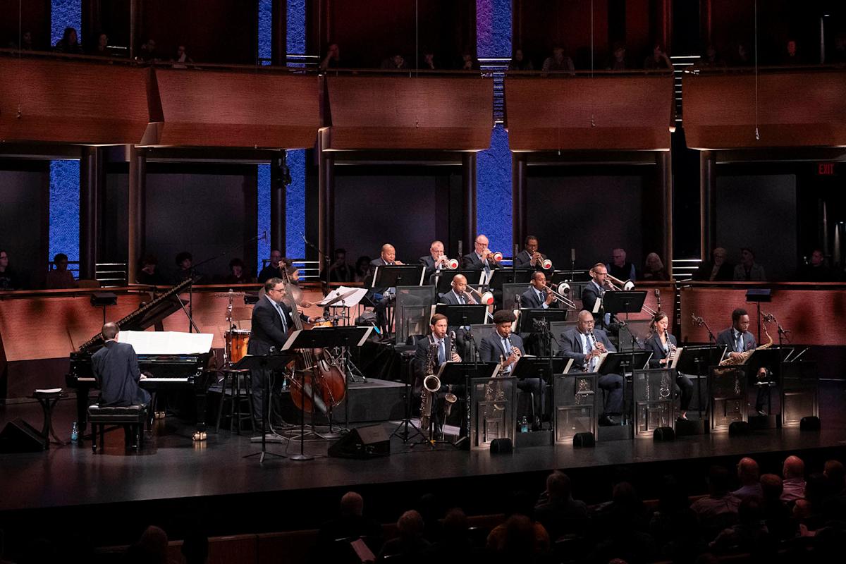 A jazz big band performs on stage with saxophones, trumpets, trombones, double bass, drums, and a piano, in front of an audience in a concert hall with blue-lit walls.