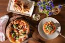 Overhead view of a table with a pizza on a wooden board, a plate of sliced salmon topped with greens, a rectangular dish with seafood, glasses of white wine, a white napkin, and a vase of colorful wildflowers.