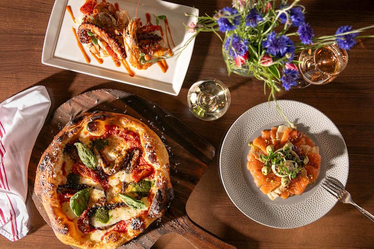Overhead view of a table with a pizza on a wooden board, a plate of sliced salmon topped with greens, a rectangular dish with seafood, glasses of white wine, a white napkin, and a vase of colorful wildflowers.