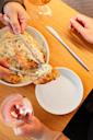 Two people share a meal of creamy pasta with fried shrimp, serving food onto a white plate, accompanied by glasses of rosé wine on a wooden table.