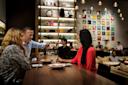 A man pours wine for a woman at a stylish, modern restaurant bar while others dine and converse in the background. The space is warmly lit, with shelves and colorful artwork decorating the walls.