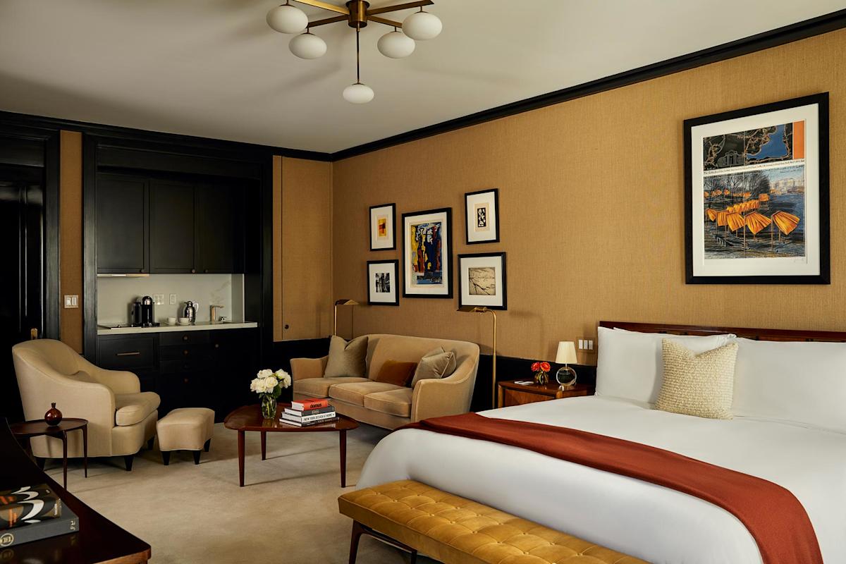 A stylish hotel room with a large bed, beige walls, modern art, a tan sofa, armchair, wooden table with books and flowers, a cushioned bench, and a small kitchenette area with a coffee maker.