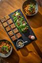 A slate platter with neatly arranged rectangular pieces of savory dish, greens with soft-boiled eggs and sundried tomatoes, flanked by two bowls of noodles and small containers of sauce on a wooden table.