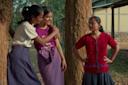 Three girls stand outdoors by trees, two of them smiling and talking closely while the third girl, wearing a red shirt, stands nearby with her hands on her hips, looking at them.