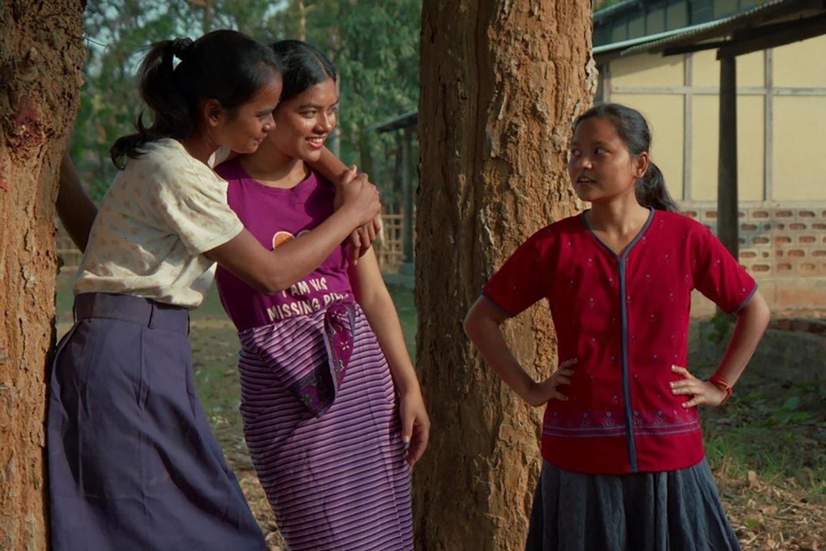 Three girls stand outdoors by trees, two of them smiling and talking closely while the third girl, wearing a red shirt, stands nearby with her hands on her hips, looking at them.