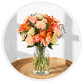 A glass vase filled with an orange and peach-toned bouquet of roses, alstroemeria, and freesia, placed on a woven table mat.