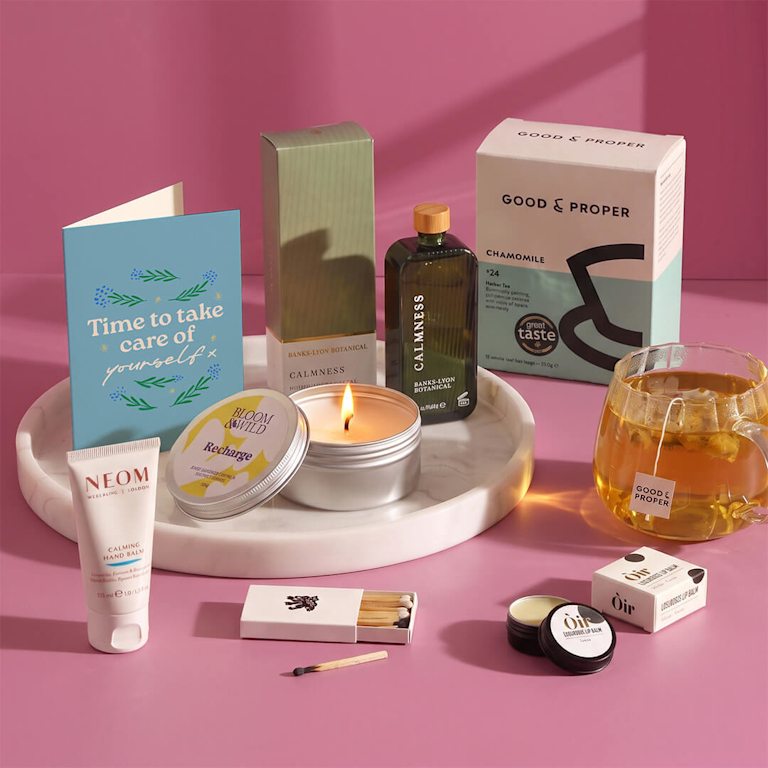 Self-care tray set with NEOM calming hand balm, soothing chamomile tea, a lit scented candle, lip balm, and a card reading "Time to take care of yourself".