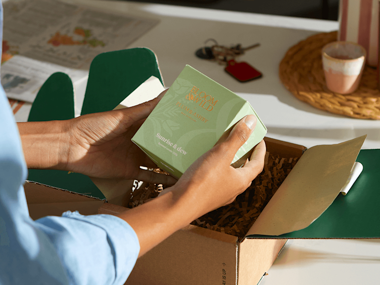 The sunrise and Dew candle being lifted out of it's Bloom & Wild packaging by 2 hands with blue sleeves.