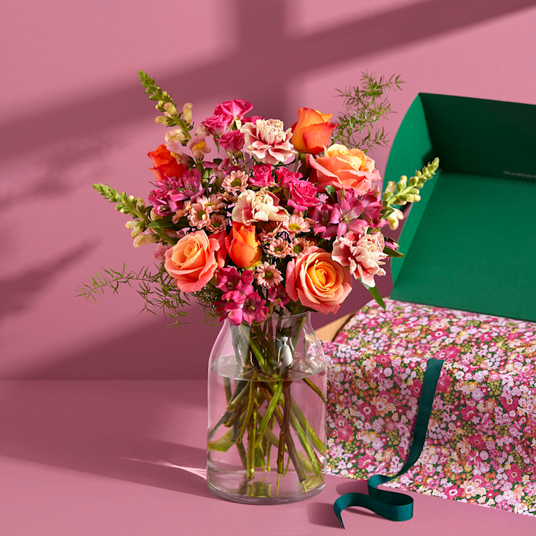 A colourful bouquet of orange, pink and yellow roses and carnations in a clear vase sits on a table laid for dinner.