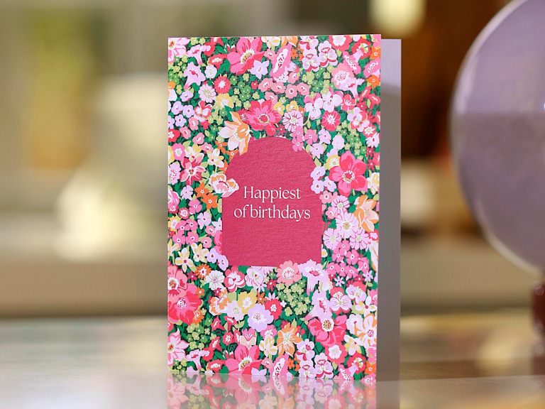 A birthday card with a floral design and the words "Happiest of birthdays" stands on a table set for dinner.