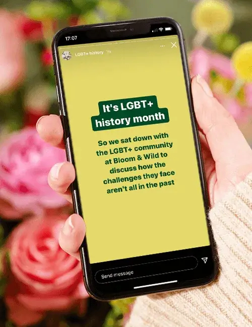 A hand holds a phone in front of a floral background. On the phone is some text talking about LGBT+ History Month.