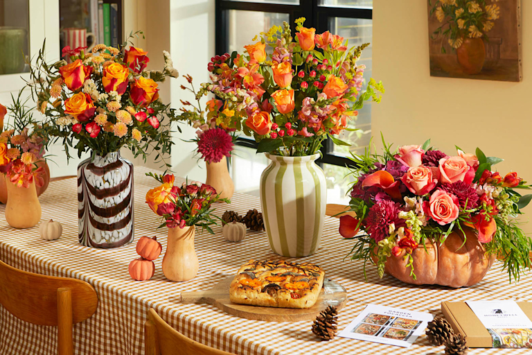 Three autumnal Bloom & Wild bouquets – the Aut and The Orla – on a warmly lit dining table next to a variety of pumpkins and squash in different colours.