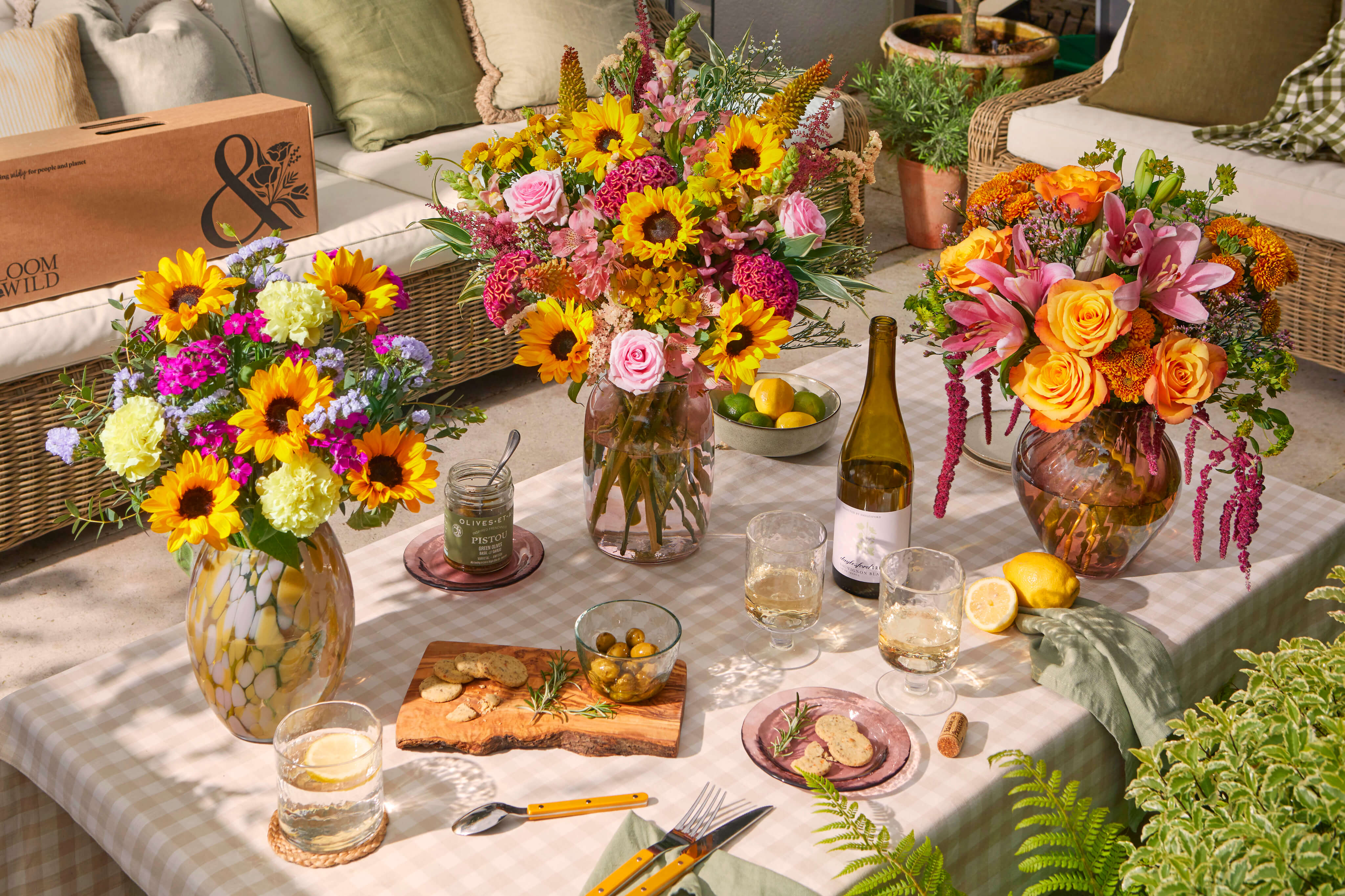 Outdoor table set for a late summer picnic with three vibrant Bloom & Wild bouquets of sunflowers, roses, and celosia in glass vases, alongside olives, crackers, wine, lemons, and gingham linens on a sunlit patio.
