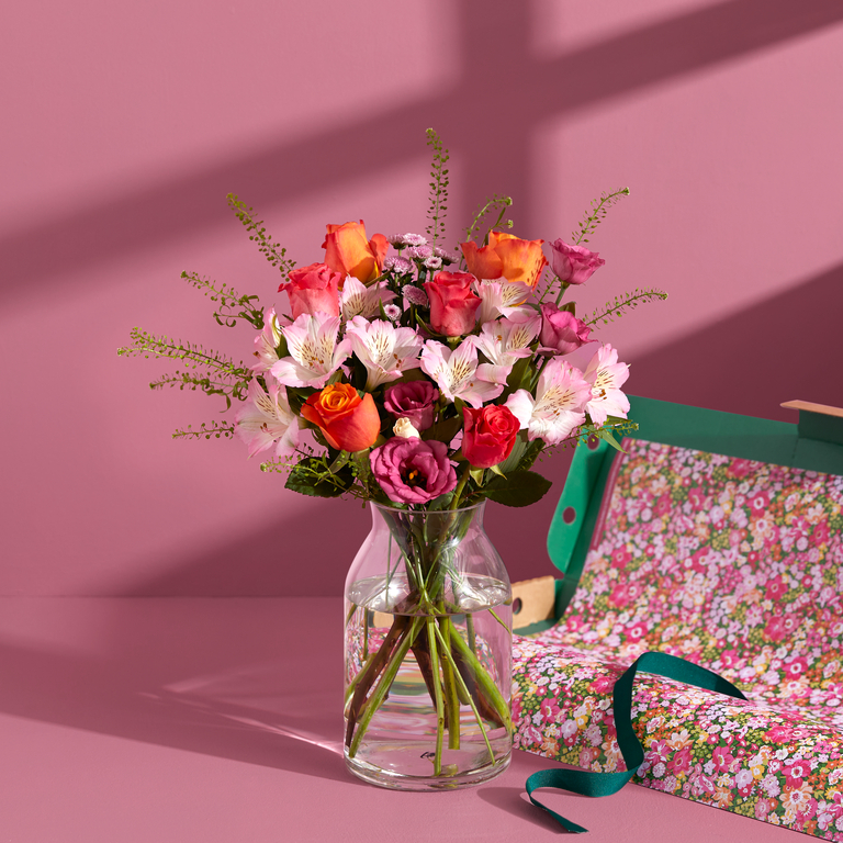 a vibrant bouquet of red and pink flowers, including roses, carnations, and chrysanthemums. The bouquet is arranged in a modern pink vase. The setting is a potting table, with gardening tools and terracotta pots in the background.