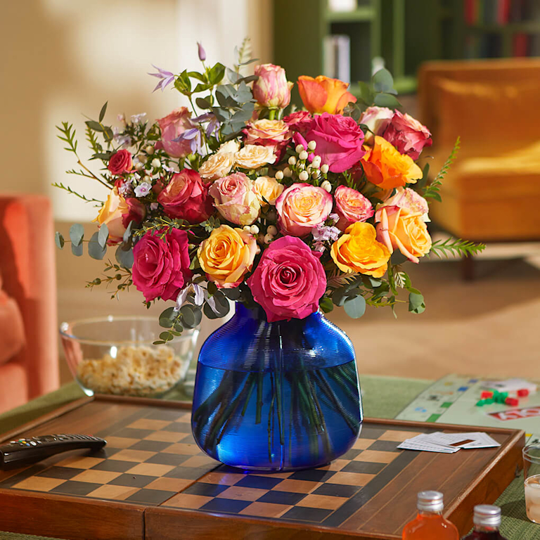 An abundant bouquet of pink, orange, and peach roses, purple limonium, and eucalyptus in a blue vase on a checkerboard.