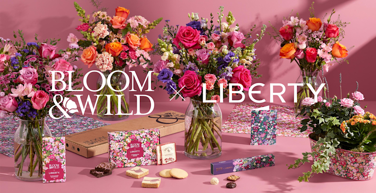 Three floral bouquets in glass vases, alongside chocolates and biscuits, sit on a pink surface with the text "BLOOM & WILD x LIBERTY".