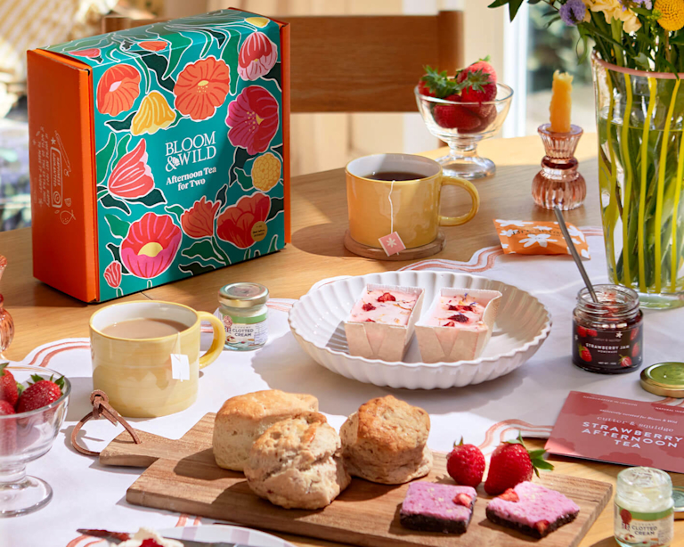 A delightful afternoon tea spread. A Bloom & Wild branded box, a yellow mug, and a vase of flowers are arranged on a wooden table. Scones, strawberries, and sweet treats sit on a white doily.