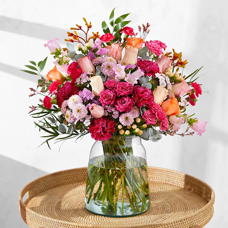 A large, textured bouquet of peach roses, magenta carnations, and lilac chrysanthemums in a clear glass jar on a woven tray. Soft sunlight hits the blooms, highlighting their delicate petals and fresh, floral aroma.