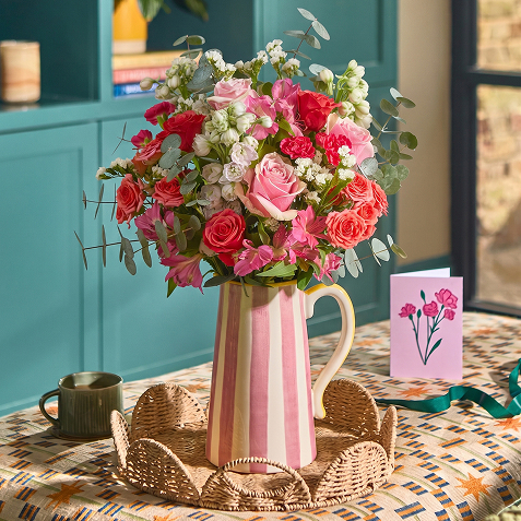 A pink and cream striped ceramic jug filled with a bouquet of pink roses, carnations, and white gypsophila, on a woven tray beside a mug and a pink greeting card with a floral illustration.