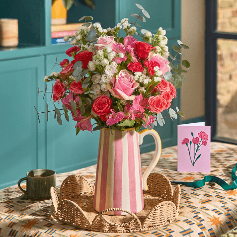A pink and cream striped ceramic jug filled with a bouquet of pink roses, carnations, and white gypsophila, on a woven tray beside a mug and a pink greeting card with a floral illustration.