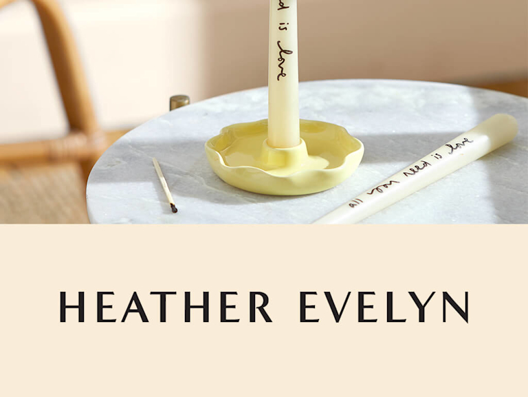 A single tapered candlestick, which is lit, in a scallop shaped ceramic candleholder. A handwritten message on the candle says 'all you need is love'