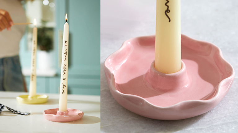 Two tapered ivory candlesticks with the message 'all you need is love' imprinted on them. One sits in a yellow scallop-edged candlestick holder, and the other in a pastel pink one.