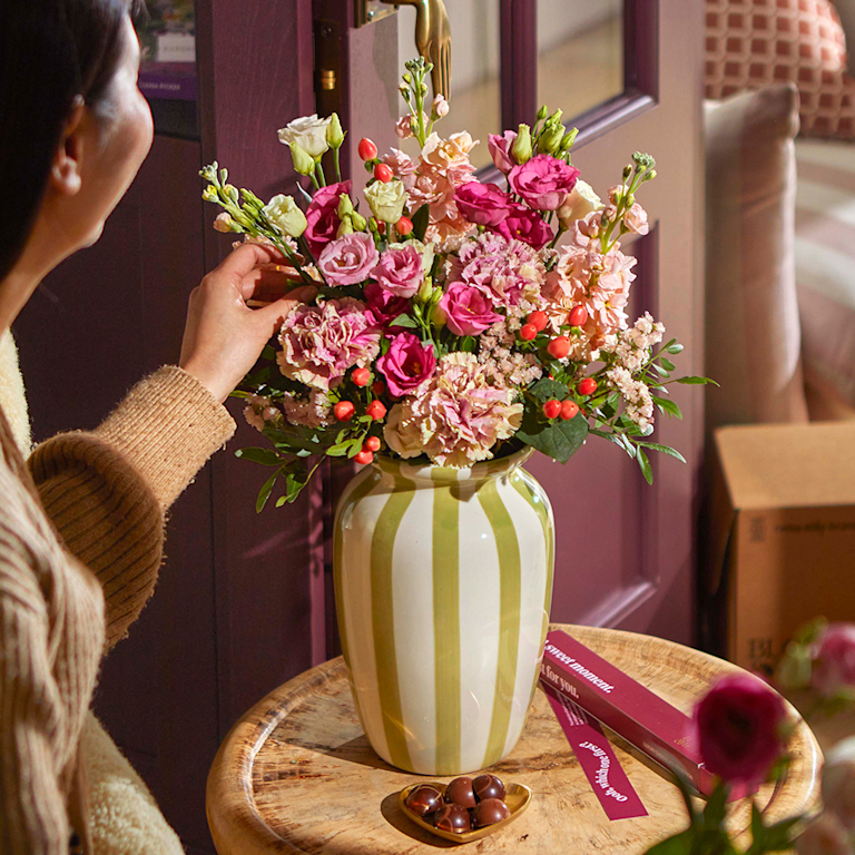 A person arranges a lush bouquet of pink lisianthus, peach stocks, and red berries in a green-striped vase. Beside a tray of chocolates, this scene feels cosy and sweet-scented, capturing a mindful moment of floral styling.