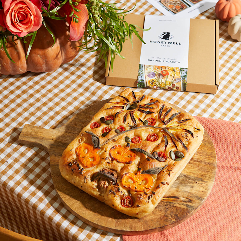 A rustic board holds a golden focaccia bread, topped with intricate pumpkin and spiderweb designs made from tomatoes, pumpkin seeds, and red onions. Behind it is the Honeywell Bakes bread kit.
