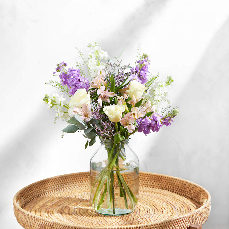 A dreamy bouquet of fragrant lilac and white stocks, cream roses, and delicate pink alstroemeria in a glass jar. Soft, ruffled textures and a sweet, spring scent. Set on a woven rattan tray against a bright, sun-dappled marble wall