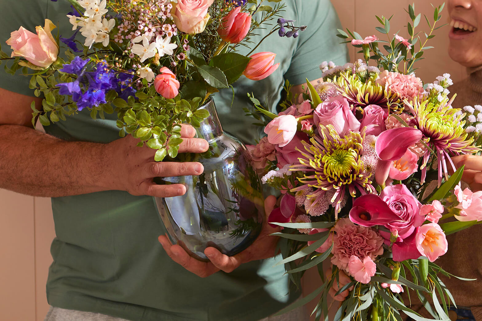Two people are stood, holding abundant vases of vividly-coloured flowers.