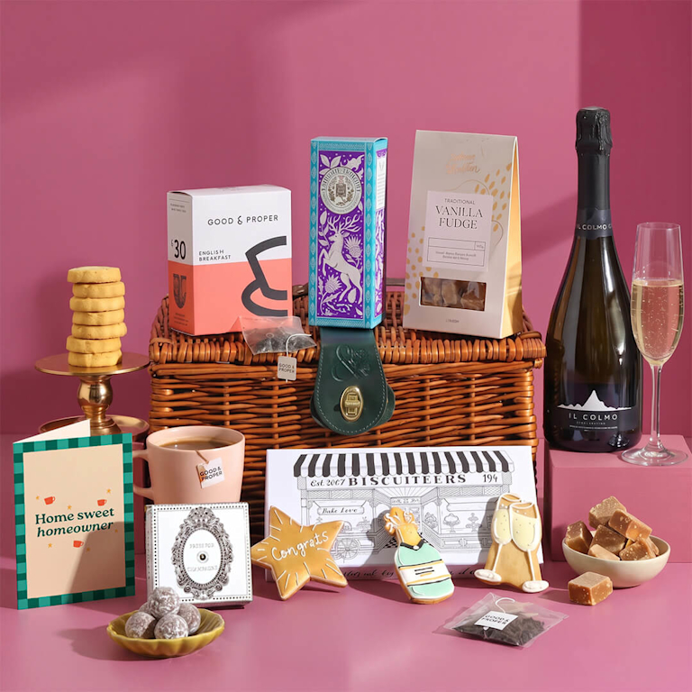 Abundant wicker hamper with prosecco, Good & Proper tea, iced biscuits, fudge, a ceramic mug, and a celebratory card reading "Home sweet homeowner" on pink.