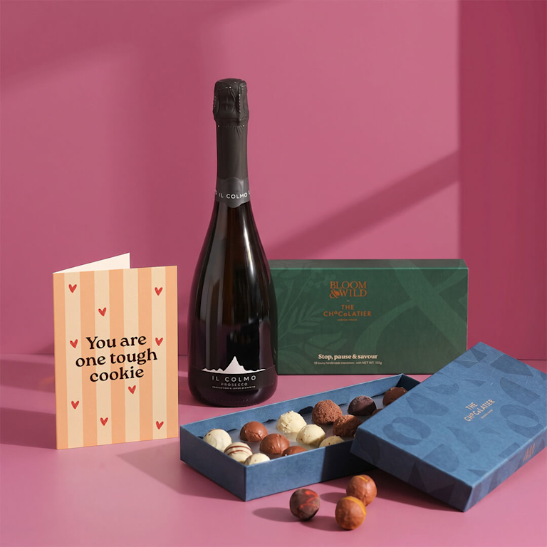 Supportive celebration set featuring Il Colmo prosecco, an open box of luxury chocolate truffles, and a card reading "You are one tough cookie" on pink.