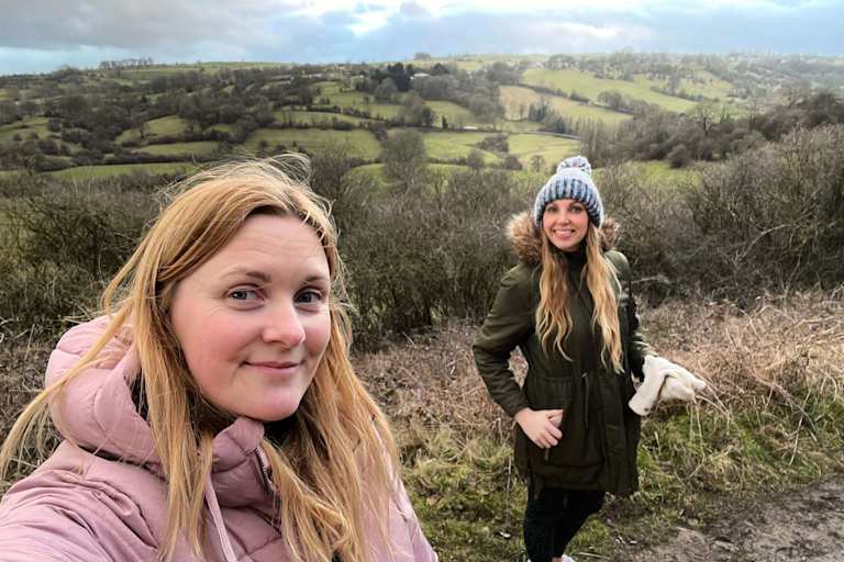 Amanda and Beth are two white women wearing coats and hats, out on a walk in the hills and smile at the camera.