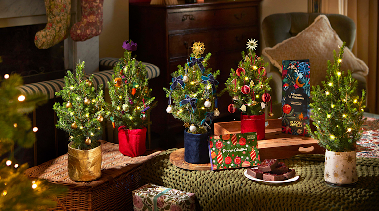 Mini Christmas trees are dotted around a living room. In between them are festive snacks, lit candles and an embroidered stocking.