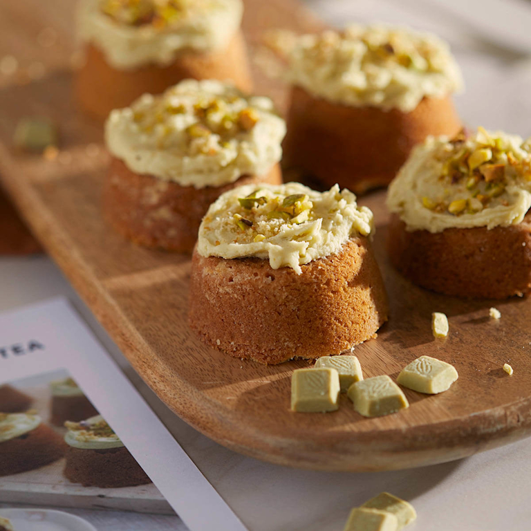 Five matcha and pistachio cakes sit on a wooden serving board. The cakes are topped with a generous swirl of pale green matcha frosting and sprinkled with chopped pistachios.